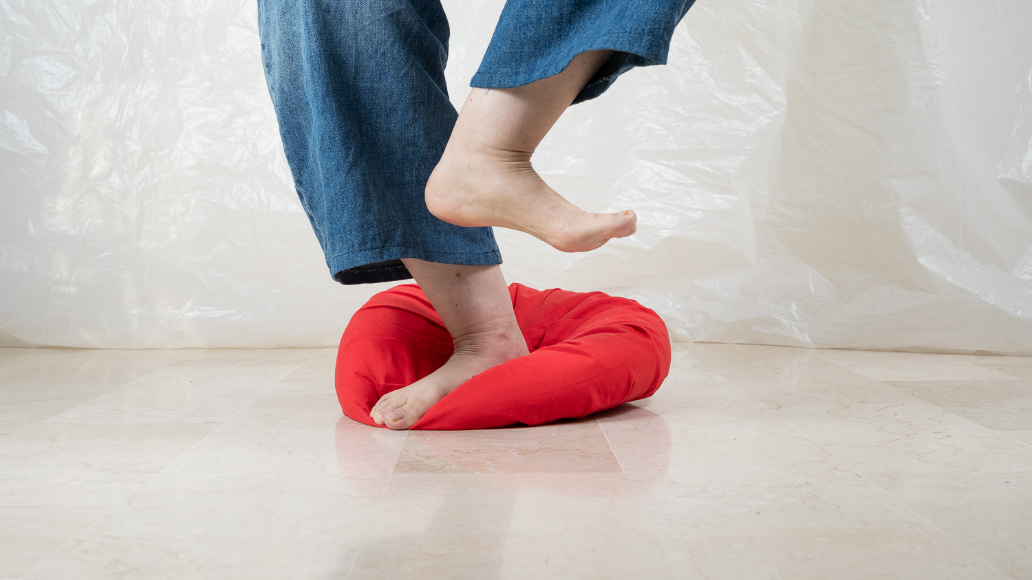 Pieds nus d'une personne en jean bleu en mouvement : un pied enfoncé dans un coussin rond rouge posé sur un sol en marbre clair, l'autre pied levé dans les airs, devant un fond de tissu plastique blanc froissé.