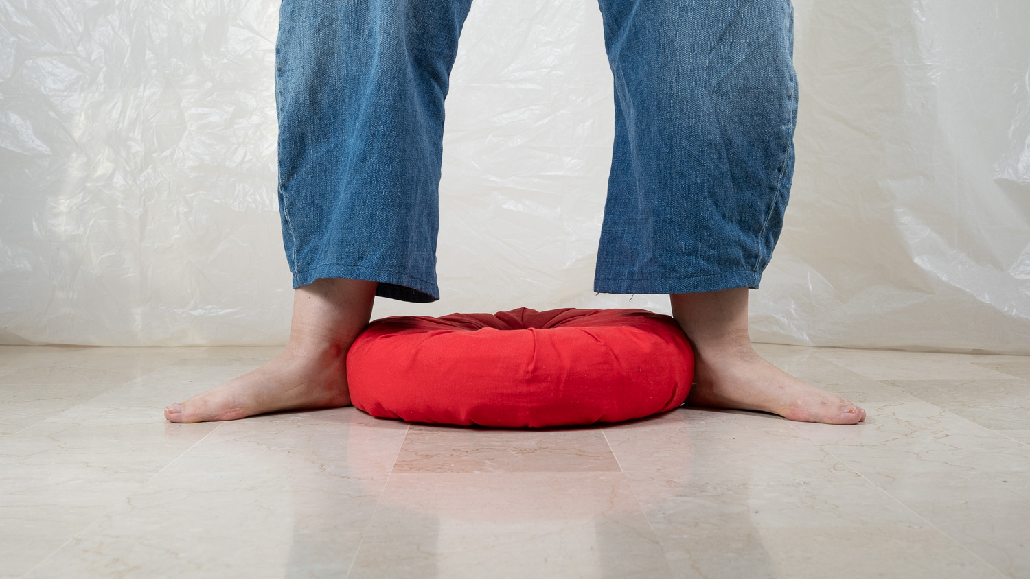 Pieds nus d'une personne en jean bleu posés sur le sol, de chaque côté d'un petit coussin rond rouge, sur un sol en marbre clair, devant un fond de tissu plastique blanc froissé.